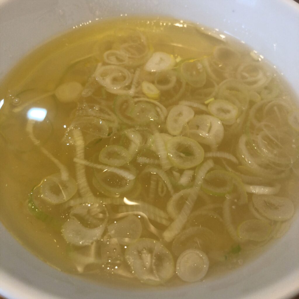 ぬまちゃんラーメンの「つけめん澄み」の鶏清湯スープ。透き通った黄金色のスープに輪切りの白ねぎが浮かび、上品な香りと旨味を感じさせる。