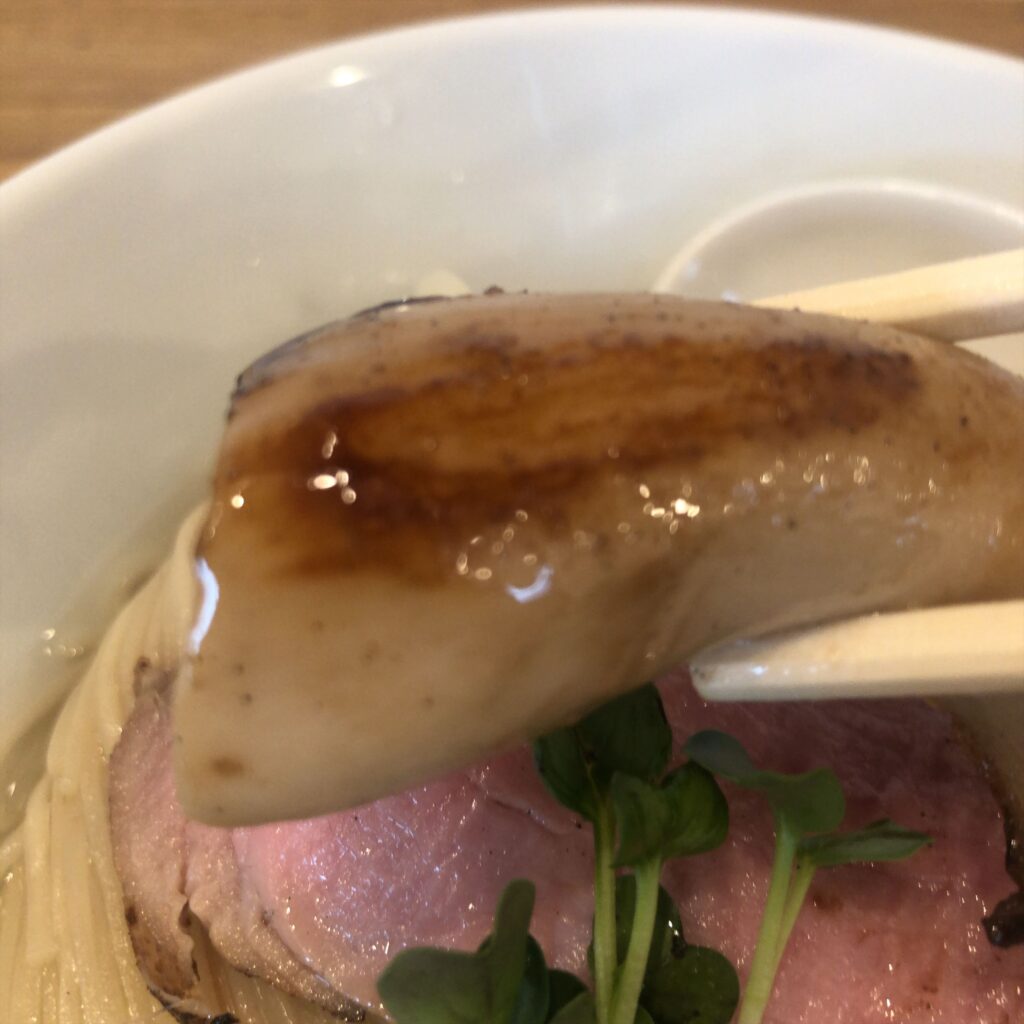 ぬまちゃんラーメンの「つけめん澄み」にトッピングされたソテーエリンギ。箸で持ち上げた様子で、香ばしい焼き目とツヤのある表面が印象的。
