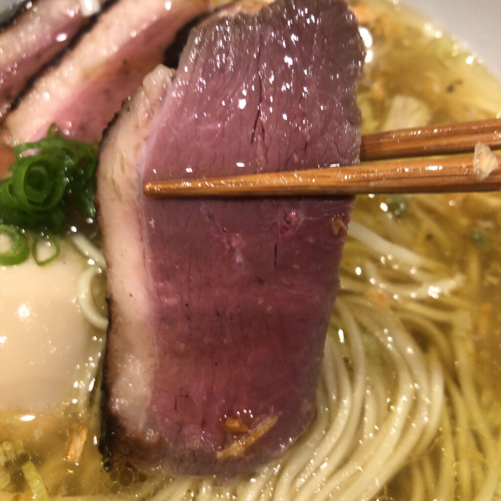 箸で持ち上げた厚切りの鴨チャーシュー。スープと麺が背景に写る。
