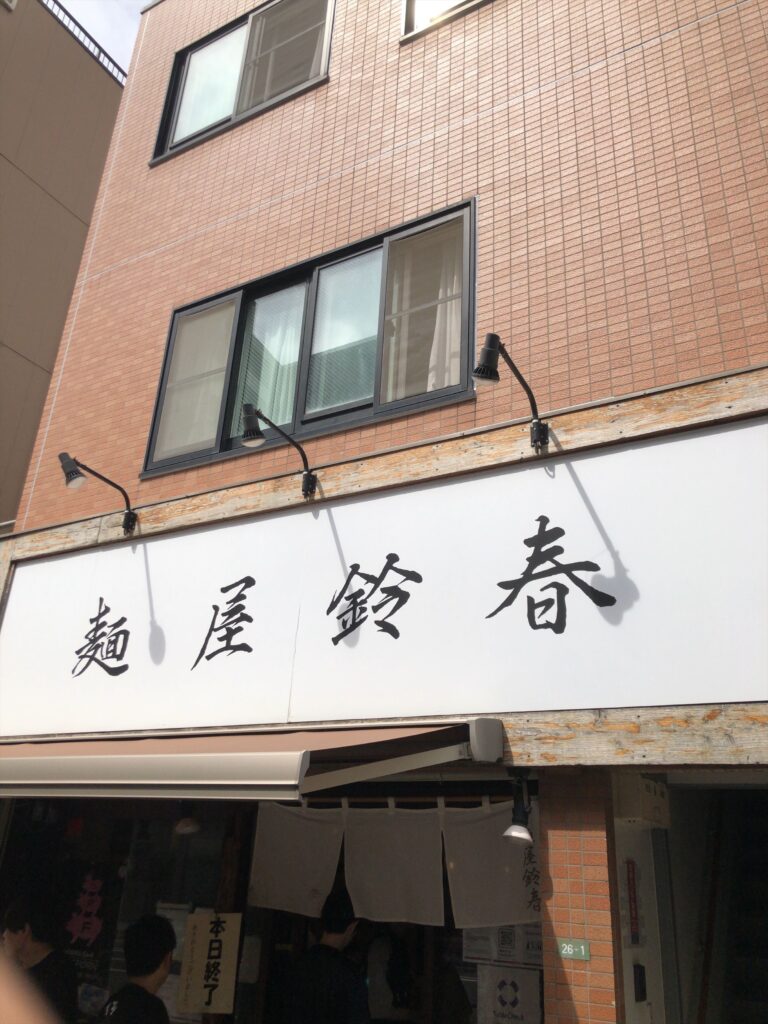 「本郷三丁目の人気店『麺屋 鈴春』の外観。白い看板に大きく店名が書かれ、暖簾の前には開店前から行列ができている様子。」