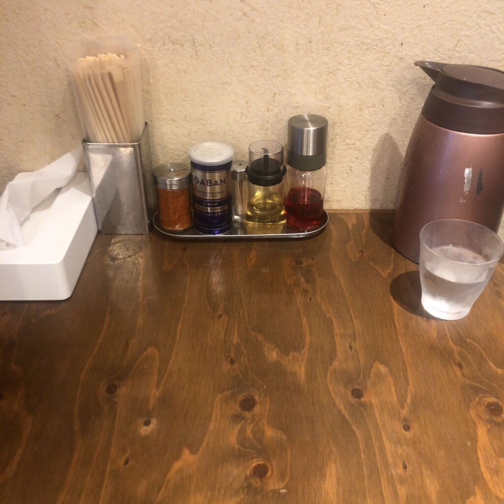 麺屋 鈴春のカウンター卓上。箸立て、ティッシュ、一味唐辛子、GABANブラックペッパー、酢、ラー油、水ポットとグラスが整然と並んでいる様子。